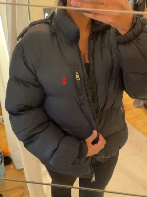 Svart pufferjacka från Ralph Lauren - Säljer en svart pufferjacka från Ralph Lauren för både tjej och kille. Har en  huva och dragkedja framtill. Jackan har en bröstficka med dragkedja och den klassiska röda Polo-loggan broderad på bröstet.