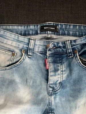 Ljusblå jeans från Dsquared2 - Säljer ett par ljusblå jeans från Dsquared2 med klassisk femficksmodell och snyggt tvättad finish. Jeansen har raka ben och detaljer som röd logotagg vid gylfen. Perfekta för en avslappnad och trendig look.