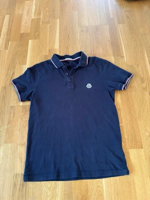 Marinblå pikétröja från Moncler - Klassisk marinblå pikétröja från Moncler med broderad logga på bröstet. Tröjan har krage med knappar och diskreta ränder i vitt och rött på krage och ärmslut. Perfekt för en stilren look.