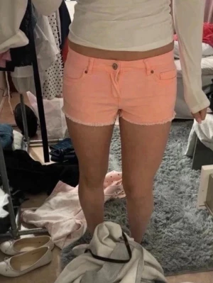 Aprikos jeansshorts med fransig kant - Defekter syns på sista bilden. Säljer ett par ljusrosa jeansshorts med fransig kant nertill. Shortsen har klassisk femficksdesign, knapp och dragkedja framtill. Perfekta för sommaren och ger en fräsch vibe till din outfit.