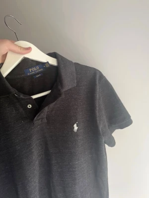 Svart pikétröja från Polo Ralph Lauren - Klassisk svart pikétröja från Polo Ralph Lauren med vit broderad logga på bröstet. Tröjan har korta ärmar, krage och knappar framtill. Perfekt för en stilren och avslappnad look.