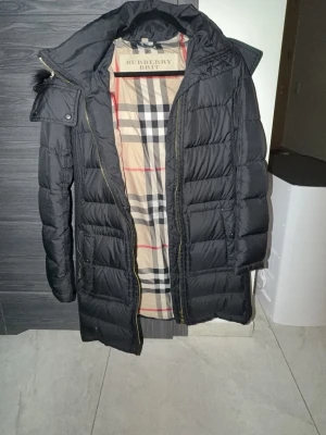 Svart dunjacka från Burberry Brit - Säljer en svart lång dunjacka från Burberry Brit med klassiskt rutigt foder. Jackan har huva, dragkedja och tryckknappar samt två stora fickor framtill. Perfekt för kalla vinterdagar och riktigt snygg streetstil.