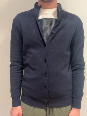 Mörkblå cardigan med ståkrage från LA CHEMISÉ - Mörkblå cardigan med ståkrage och knäppning framtill. Tröjan har långa ärmar och ribbade muddar vid ärmslut och nederkant. Enkel och stilren design, perfekt för lager-på-lager. Materialet ser ut att vara mjukt och stickat. Hör av er gällande funderingar 