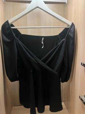 Svart blus med puffärm - Trendig svart blus med puffiga trekvartsärmar och omlott-detalj framtill. Blusen har en lätt ribbad struktur och är perfekt för dig som gillar en stilren men ändå cool look. Materialet känns mjukt och följsamt mot huden. Väldigt bekväm. Storlek 36/38
