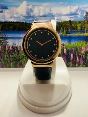 Stilren Mörklilia  klocka med gulddetaljer - Snygg klocka med mörklila  urtavla och guldfärgat boett. Urtavlan har minimalistiska markeringar med små stenar och matchande rosé guldfärgade visare. Armbandet är mörklila och har en klassisk design. Perfekt accessoar för en trendig look.Nytt batteri, säljes i befintligt skick enligt bilderna!