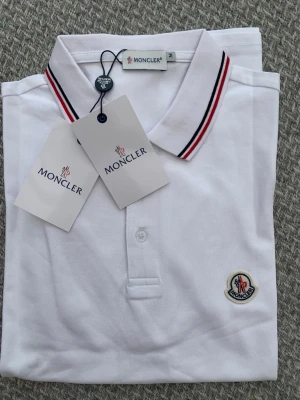 Vit pikétröja från Moncler - Klassisk vit pikétröja från Moncler med broderad logga på bröstet. Kragen har snygga ränder i rött och svart för en sportig look. Tröjan har knappar framtill och är tillverkad i bomull för en bekväm känsla.