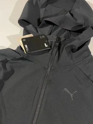Svart Puma träningsjacka/ hoodie  - Denna personliga favorit tröja med huva och dragkedja framtill från Puma är verkligen top klass. Stilren tech med mattsvart färg. Luvtröjan har två sidofickor med dragkedja och är tillverkad i ett lätt och funktionellt material med DryCELL-teknologi som transporterar bort fukt. Perfekt för träning utomhus eller gymmet.