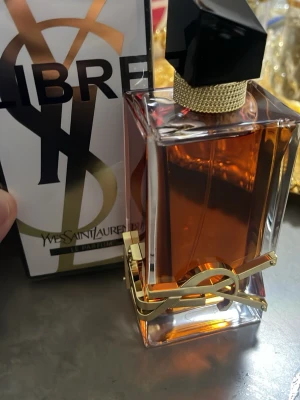 Yves Saint Laurent Libre Eau de Parfum - Yves Saint Laurent Libre Eau de Parfum med elegant glasflaska, guldiga detaljer och svart asymmetrisk kork. Parfymen har en varm bärnstensfärgad vätska och lyxig design med YSL-logga i metall runt flaskan. Perfekt för dig som gillar exklusiva dofter.