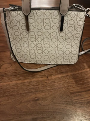 Beige axelväska från Calvin Klein - Snygg beige axelväska från Calvin Klein med CK-logga över hela väskan. Väskan har justerbar axelrem, silverfärgade detaljer och är tillverkad i konstläder. Perfekt storlek för det viktigaste och ger en clean look till din outfit.