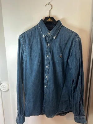 Blå jeansskjorta från Ralph Lauren - Snygg blå jeansskjorta från Ralph Lauren i slim fit-modell. Skjortan har klassisk krage, knäppning framtill och broderad logga på bröstet. Perfekt för en avslappnad men stilren look.