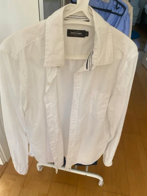 Vit skjorta från Dressmann - Klassisk vit skjorta från Dressmann med lång ärm och bröstficka. Skjortan har en stilren design med knappar framtill och en mjuk krage. Perfekt för en clean och enkel look.