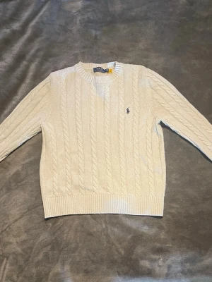  Ralph Lauren Tröja - Kabelstickad vit tröja från Polo Ralph Lauren med rund halsringning och broderad logga på bröstet. Tröjan har långa ärmar och ribbade muddar vid ärmslut och nederkant. Perfekt för en stilren och klassisk look.