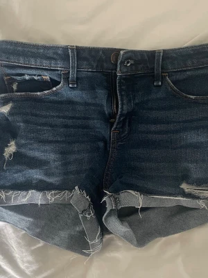 Mörkblå jeansshorts med slitningar - Snygga mörkblå jeansshorts med råa kanter och slitna detaljer. Klassisk femficksmodell med dragkedja och knapp framtill. Perfekta för varma dagar och en avslappnad stil.