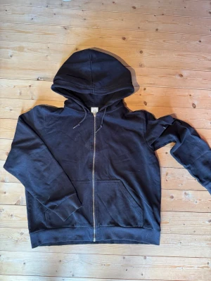 Svart zip hoodie  - Klassisk svart hoodie från H&M Basics med dragkedja framtill och två stora fickor. Tillverkad i mjukt bomullsmaterial och har en justerbar huva med snören. Perfekt för en avslappnad och enkel stil.