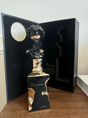 Lattafa His Confession Parfym 100ml - Lattafa His Confession är en lyxig parfym i en svart och guld flaska med unik design, där korken föreställer en klassisk byst. Flaskan rymmer 100 ml och levereras i en elegant svart förpackning med gulddetaljer. Perfekt för dig som gillar exklusiva dofter och snygg inredning.
