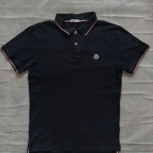 Svart pikétröja från Moncler - Klassisk svart pikétröja från Moncler med korta ärmar och knappslå framtill. Kragen och ärmsluten har snygga rödvita ränder som ger en sportig touch. Moncler-logga broderad på bröstet. Perfekt för en stilren och avslappnad look.