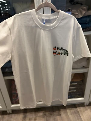 Vit Off-White x KAWS t-shirt - Vit t-shirt från Off-White i samarbete med KAWS. Framsidan har en liten grafisk detalj på bröstet och baksidan har en stor färgglad print med KAWS-figur och Off-White-logga. Klassisk passform och rund hals. Perfekt statement-plagg för streetwear.