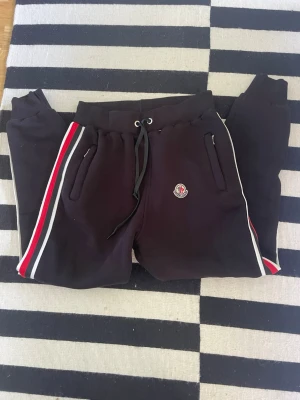 Svarta Moncler mjukisbyxor med ränder - Säljer ett par svarta mjukisbyxor från Moncler med vita och röda ränder längs sidorna. Byxorna har dragsko i midjan, muddar vid bensluten och två fickor med dragkedja. Moncler-logga på vänster ben. Perfekta för en chill och sportig look.