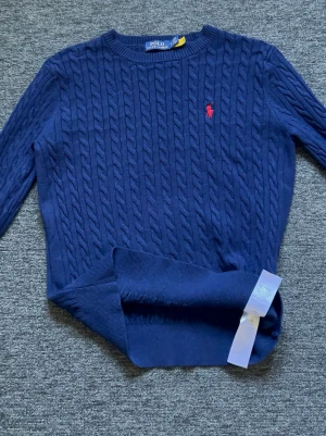 Mörkblå kabelstickad tröja Polo Ralph Lauren - Kabelstickad mörkblå tröja från Polo Ralph Lauren med rund halsringning och röd broderad logga på bröstet. Tröjan har långa ärmar och ribbade muddar. Perfekt för dig som gillar klassisk och stilren look.