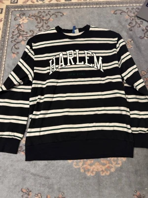 Svartvit randig sweatshirt Harlem - Cool svart och vit randig sweatshirt med texten HARLEM New York på bröstet. Tröjan har rund halsringning och långa ärmar. Perfekt för en avslappnad streetwear-look.