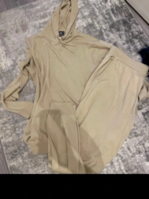 Beige mjukisdress med huva - Säljer en beige loungewear-set med hoodie och matchande byxor. Hoodien har dragsko i huvan och byxorna har resår i midjan för extra komfort. Perfekt för chill dagar hemma. Materialet känns mjukt och skönt mot huden.