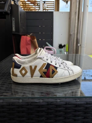 Gucci Loved vita sneakers - Unika vita sneakers från Gucci med texten 'LOVED' i guld på sidan. Skorna har klassiska Gucci-ränder i rött och blått samt metallic-detaljer i rött och blått på hälen. Tillverkade i läder med vita snören och låg profil. Perfekta för dig som vill sticka ut. Tveka inte på att höra av dig vid minsta fråga och våga skicka prisförslag💕