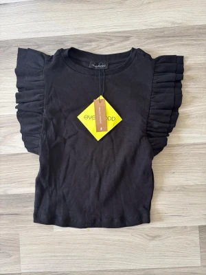 Svart topp med volangärm från Even&Odd - Trendig svart topp från Even&Odd med ribbad struktur och volangdetaljer på axlarna. Tillverkad i ekologisk bomull och elastan för en bekväm passform. Perfekt för dig som vill ha en stilren men ändå unik look.
