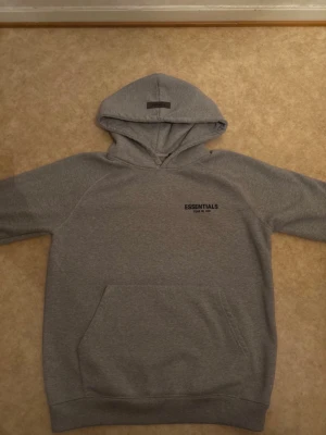 Grå Essentials Fear of God hoodie - Snygg grå hoodie från Essentials Fear of God med svart logga på bröstet. Klassisk känguruficka framtill och stor huva. Tillverkad i mjukt bomullsmaterial, perfekt för en avslappnad streetwear-look.