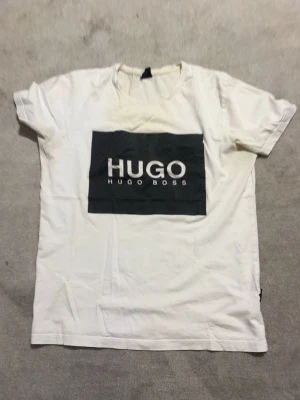 Vit t-shirt från Hugo Boss - Vit t-shirt från Hugo Boss med stor svart logga på bröstet. Klassisk passform och rund halsringning. Perfekt för en stilren och enkel look.