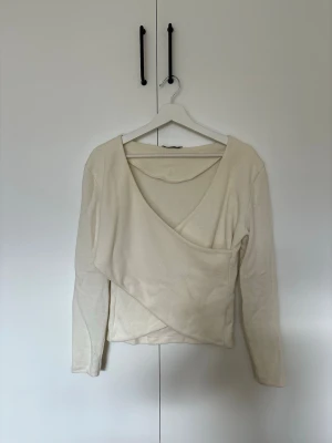 Beige omlottopp med v-ringning - Trendig beige omlottopp med lång ärm och djup v-ringning. Toppen har en snygg omlottdesign som ger en cool och avslappnad look. Perfekt att styla med jeans eller kjol för en modern vibe.