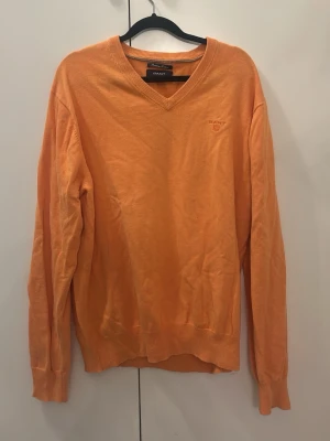 Orange stickad tröja från GANT - Snygg orange stickad tröja från GANT med v-ringning och diskret broderad logga på bröstet. Tröjan har långa ärmar och ribbade muddar vid ärmslut och nederkant. Perfekt för lager-på-lager och enkel att matcha. 