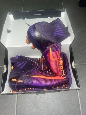 Nike Mercurial Elite lila/orange - Säljer ett par Nike Mercurial fotbollsskor i lila och orange med högt skaft och strumpdesign. Skorna är helt nya och aldrig använda, endast testade inomhus. Skorna är av sällsynt modell som ej säljs på förstahands marknaden längre. Priset är ej hugget i sten. Skopåse medföljer, ej orginal skolåda.