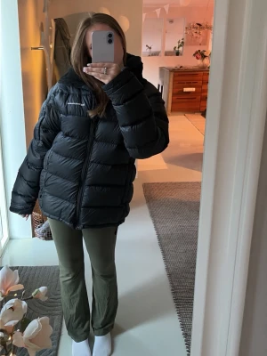 Peak Performance dunjacka - Säljer en svart pufferjacka från Peak Performance med huva och dragkedja framtill. Jackan är vadderad och har en oversized passform som ger en cool och avslappnad look. Det finns ett hål i den vänstra armhålan som är lagad med en patch, tyvärr har den släppt lite och kan behövas bytas ut annars mycket fint skick