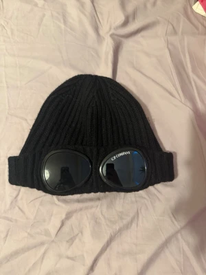 Svart gogglemössa från C.P. Company - Säljer en svart ribbstickad mössa från C.P. Company med ikoniska goggle-linser framtill. Mössan är i mjukt stickat material och har uppvikt kant. Perfekt för dig som vill sticka ut med en streetwear-inspirerad look.