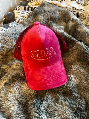 Rosa Von Dutch keps med mesh - Säljer en snygg röd keps från Von Dutch med glittrande stenar på loggan framtill. Kepsen har klassisk trucker-modell med mesh baktill och justerbar rem. Materialet framtill är i mjuk mockaimitation som ger en lyxig känsla. Säljer för jag inte använder den. 