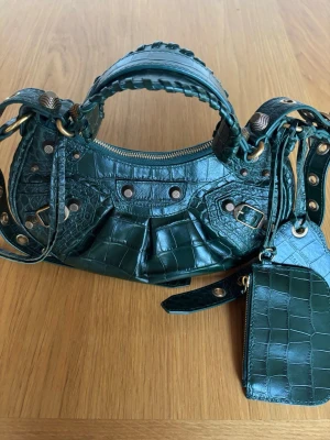 Grön Balenciaga väska - Tryck inte på köp! Möts helst upp om du bor/har vägarna förbi i Stockholm. Intressekoll på min Balenciaga väska i modellen le cagole xs i krokopräglat läder med guldfärgade metalldetaljer. Väskan har avtagbar plånbok och spegel. Kvitto och dustbag finns. Väskan är väl omhändertagen och i mycket fint skick. 