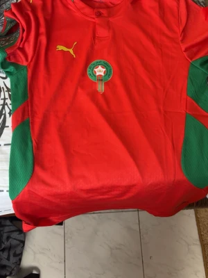 Marocko fotbollströja Puma röd/grön - Säljer en officiell Marocko fotbollströja från Puma i rött med gröna detaljer på sidorna. Tröjan har korta ärmar, knapp vid halsen och Marockos landslagsemblem på bröstet. Tillverkad i lätt och ventilerande material, perfekt för match eller träning.