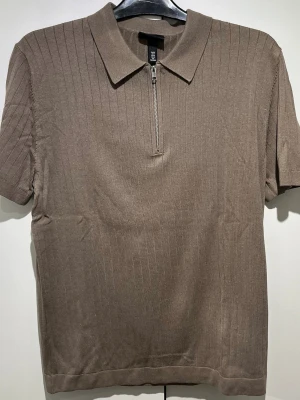 Brun ribbad polotopp med dragkedja - HM - Säljer en brun ribbad polotopp från H&M med korta ärmar och en snygg dragkedja framtill. Toppen har klassisk krage och är tillverkad i mjukt material som känns skönt mot huden. Perfekt för dig som gillar stilrena och enkla plagg.