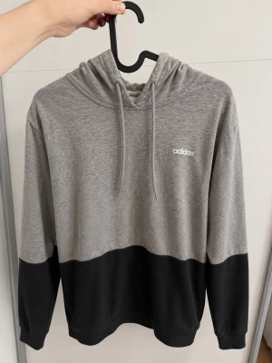 Grå och svart hoodie från Adidas - Snygg tvåfärgad hoodie från Adidas i grått och svart. Hoodien har dragsko i huvan och en liten Adidas-logga på bröstet. Tillverkad i mjukt och bekvämt material, perfekt för en avslappnad stil. Passar dig som gillar sportiga och stilrena plagg. Storlek M men går även bra som S.
