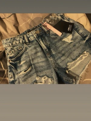 Blå denimshorts med slitningar - Säljer ett par blå denimshorts med råa kanter och slitna detaljer framtill. Klassisk femficksmodell med knapp och dragkedja. Perfekta för en avslappnad och trendig look.
