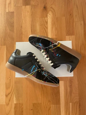 Svarta Maison Margiela "Paint Splatter" GATs - Tjena! Säljer ett par riktigt feta Maison Margiela Sneakers i den ikoniska "Paint Splatter"-modellen. Skorna är svarta med ett unikt, flerfärgat färgstänk och har den klassiska rågummisulan. Modellen är tillverkad i premiumläder och mocka. De levereras i tillhörande originalbox. Skickas välpackade så snabbt som möjligt. Hör av dig vid frågor eller om du vill ha fler bilder! ⚡️