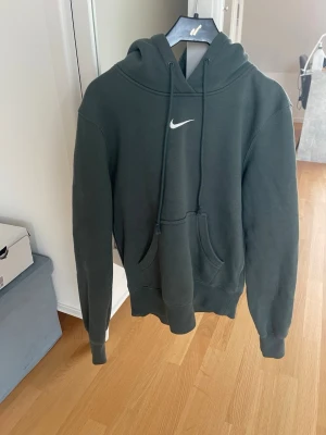 Mörkgrön Nike hoodie med huva - Snygg mörkgrön hoodie från Nike med klassisk vit logga på bröstet. Hoodien har en stor ficka framtill, justerbar dragsko i huvan och ribbade muddar vid ärmar och nederkant. Perfekt för en avslappnad och sportig stil.