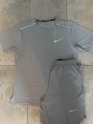 Grått träningsset från Nike - Säljer ett grått träningsset från Nike med t-shirt och shorts. Båda plaggen har diskreta vita Nike-loggor och är gjorda i ett lätt, snabbtorkande material som passar perfekt för träning. T-shirten har rund hals och korta ärmar, shortsen har resår i midjan.