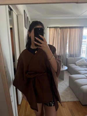 Brun cape-kappa med knytskärp - Snygg brun cape-kappa med vid passform och knytskärp i midjan. Kappan har en öppen design utan synliga knappar och är tillverkad i ett mjukt tyg som ger ett elegant fall. Perfekt för lager-på-lager och trendig look.