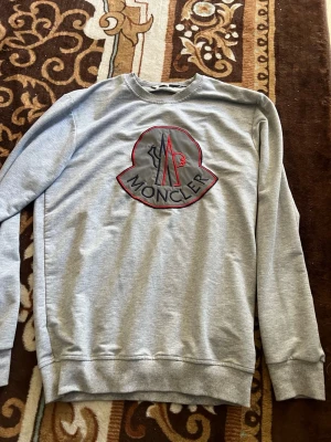 Grå sweatshirt från Moncler - Snygg grå sweatshirt från Moncler med stort broderat logotypmärke på bröstet i svart, rött och grått. Tröjan har rund halsringning, långa ärmar och ribbade muddar vid ärmslut och nederkant. Perfekt för en avslappnad och trendig look.