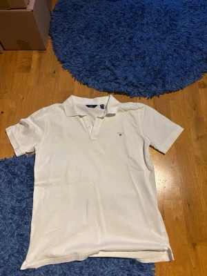Vit pikétröja från Gant - Klassisk vit pikétröja från Gant med korta ärmar och liten broderad logga på bröstet. Tröjan har krage och knappar framtill, perfekt för en clean och stilren look. Tillverkad i mjuk bomull som känns skön mot huden.
