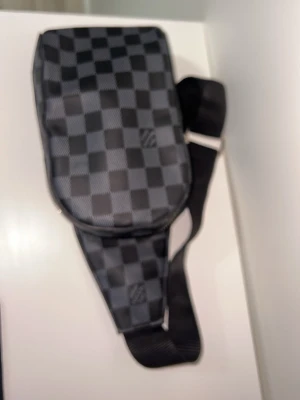 L och V - Snygg axelväska från L och V i det klassiska Damier Graphite-mönstret. Väskan är svart och grå med justerbar axelrem och har en modern, rektangulär form. Tillverkad i canvas med detaljer i läder. Perfekt för dig som vill ha en stilren och exklusiv accessoar.