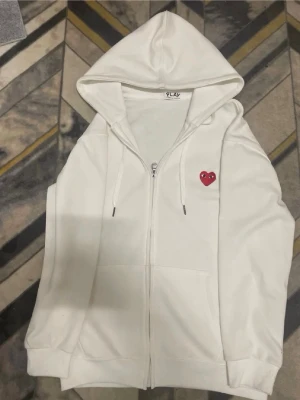 cdg kofta - Vit hoodie med dragkedja från Comme des Garçons Play. Klassisk design med huva och dragsko samt det ikoniska röda hjärtat med ögon broderat på bröstet. Tillverkad i mjukt bomullsmaterial och har två fickor framtill.