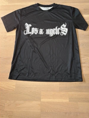 Svart Los Angeles t-shirt - Snygg svart t-shirt med stor vit text 'Los Angeles' i gotisk stil över bröstet. Klassisk passform och rund halsringning. Perfekt för dig som gillar streetwear och vill ha en enkel men stilren look.