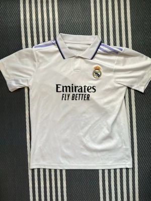 Real Madrid matchtröja Vinicius Jr - Säljer en vit Real Madrid matchtröja från Adidas med klubbmärke och 'Emirates Fly Better' tryck på bröstet. Tröjan har lila detaljer på axlarna, pikékrage och Vinicius Jr nummer 20 på ryggen. Perfekt för fotbollsfans!